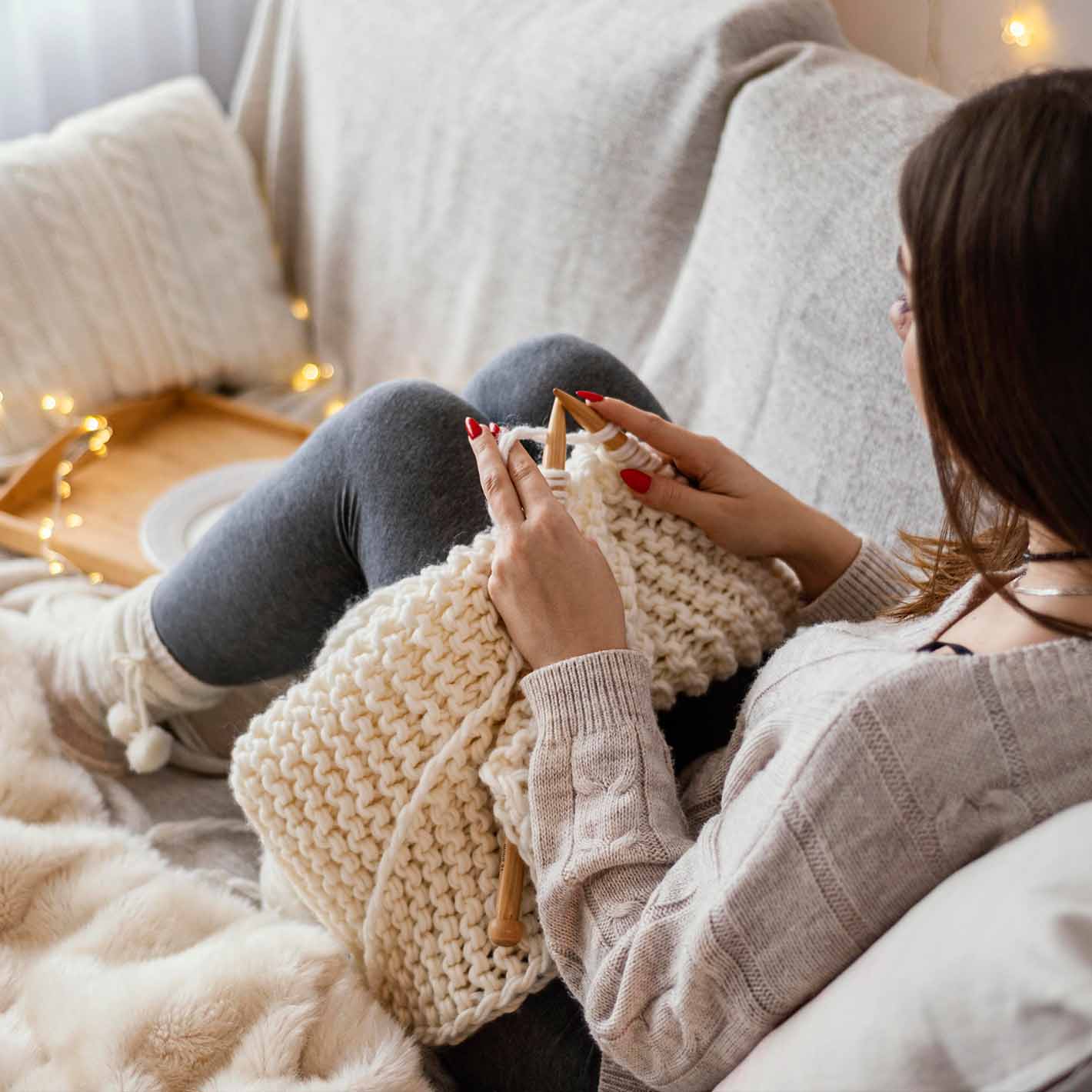 young lady knitting on couch surrounded by blankets, lights and pillow