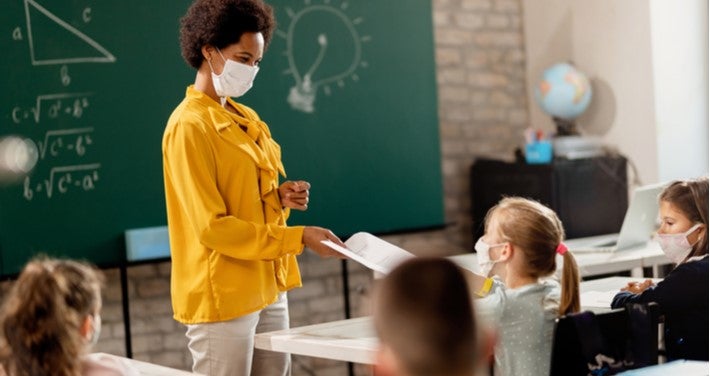 Black teacher with mask on handing student a paper in front of class