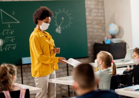 Black teacher with mask on handing student a paper in front of classroom